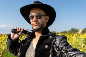 A stylish man in a black hat and sunglasses stands confidently in a vibrant sunflower field on a sunny day.